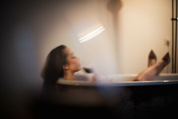 A woman with black long hair drinks red wine in a white bathtub.