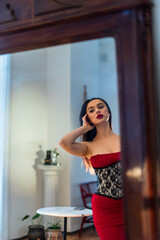 A woman in a red dress with a luxurious figure admires her reflection in the mirror. Stands on the background of a living room with white walls and furniture.