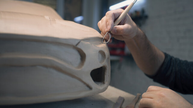Experienced car designer works on the prototype eco friendly electric model of the car using sculpting tool to carve out chunks of clay for details in the design. Hand made car sculpture made of clay.