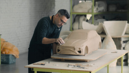 Experienced car designer with an apron works on the sculpture of the car using wooden shaping tool to smooth out the surface and create details in the design. Hand made car sculpture made of clay.