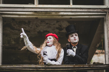 A girl with scary makeup in a white suit and a red hat shows something to a man in a black suit and a black hat. Both with glasses of wine. Halloween