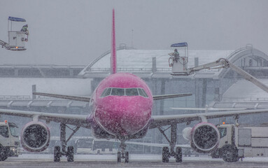 A jet passenger aircraft of pink color is serviced by deicing in winter against the background of falling snow