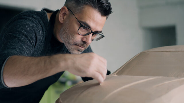 Experienced automotive designer drags across the wooden shaping tool to smooth out the surface of the car sculpture prototype. Designer with an apron works on the clay sculpture of the car.