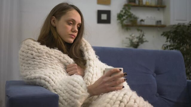 Girl sitting on couch in apartment, covered with blanket, drinks hot coffee. girl enjoys loneliness in cozy comfortable home. man drinks hot drink of coffee in winter covered with blanket.