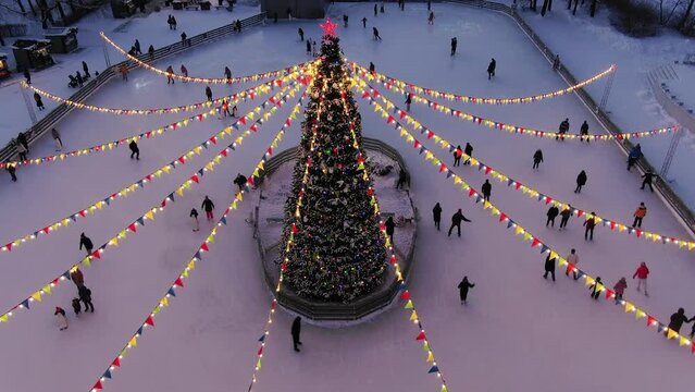 People are skating on a skating rink around a festive Christmas tree decorated with a garland and flags, aerial view