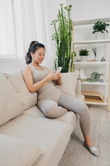 Athletic Asian woman in yoga suit blogger looking at phone in hand sitting on couch and smiling with headphones, freelance work from home, recording sports workout lifestyle body care