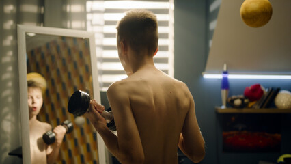 A focused school boy do dumbbells curls stands in front of the mirror in his bedroom. Nude upper...