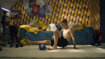 Young athlete boy uses foam roller in his room. Warm up sports training. School sports team soccer player. Stretching muscles. Workout at home.