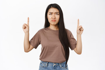 Disappointed asian woman pointing, looking up with sad, regret face, showing smth bad, standing over white background