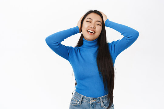 Portrait of beautiful asian woman with long, natural dark hair, touching her hairstyle, laughing and smiling, standing over white background