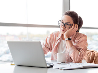 Frowned woman in eyeglasses works with laptop and hand written notes in notepad. Modern office with panoramic windows at co-working center. Workplace for freelancers or students in business center.