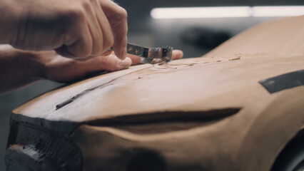 Close-up shot of male automotive designer working on futuristic car model from plasticine clay with rake. Future design of new generation electric car. Work in modern car design studio.