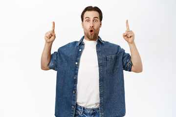 Portrait of impressed guy shows smth awesome up, pointing on top, makes wow face, stands over white background © Cookie Studio
