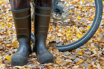 With dry feet on the bike through autumn. Cyclist stands in rubber boots in colorful foliage in...
