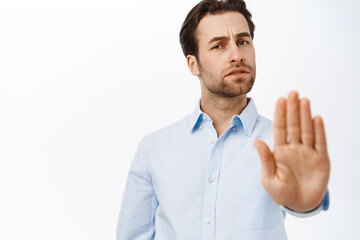 Portrait of businessman showing stop sign, disapprove smth, extends his palm in rejection, stands over white studio background