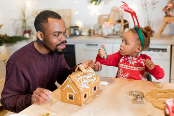African American father with daughter  using royal icing to create decorating a gingerbread house together at home. Fun Christmas activities for kids