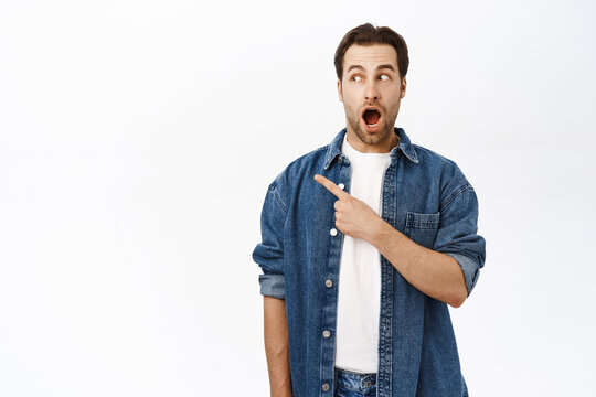 Image of handsome young man pointing finger left, saying wow, looking impressed at advertisement, standing in casual outfit over white background
