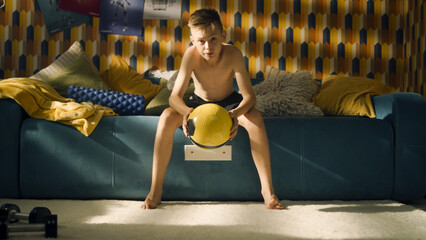 A teenage boy sits on a couch with a football in his hands. Shirtless young athlete boy. Focused for an upcoming soccer match. School sports team.