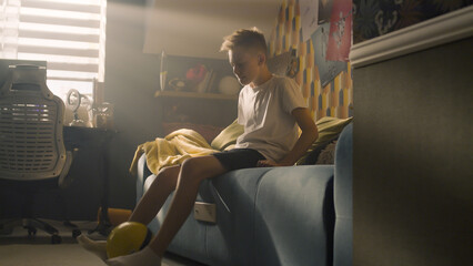A teenager boy plays football while sitting on the bed alone in his room. Balancing the ball on his finger. Performing tricks. Sports, hobbies concept. © Framestock