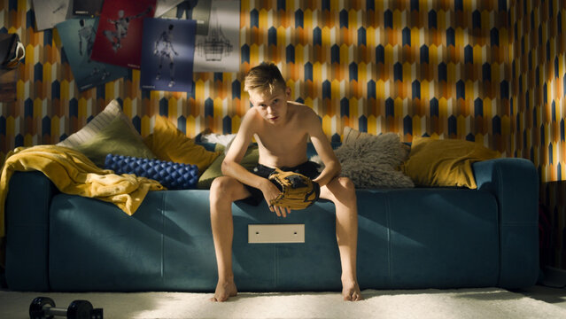 A teenage boy sits on a couch with a football in his hands. Shirtless young athlete boy. Focused for an upcoming soccer match. School sports team.