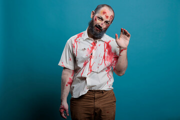 Dangerous zombie waving hello at camera, doing salute gesture over blue background. Creepy apocalyptic evil monster with bloody scars and wounds, brain eating aggressive corpse.