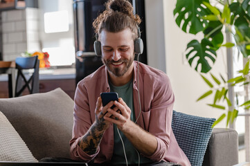 Young cheerful man with a sleeve tattoo using a laptop computer and smartphone listening to music...