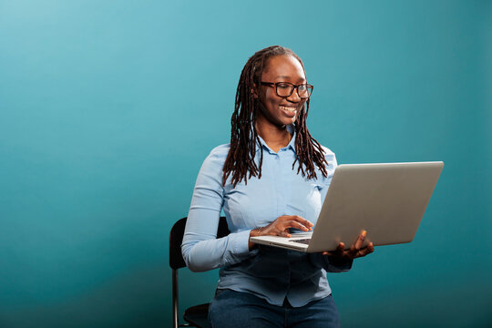 Joyful pretty lady with handheld modern computer standing on blue background while surfing browser. Cheerful happy woman having laptop and smiling heartily while browsing webpages on internet