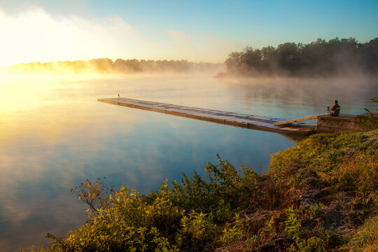 Man on wooden boardwalk pier photographing peaceful foggy lake at sunrise