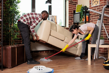 Modern wife mopping under sofa while husband lifts it, multiracial couple doing spring cleaning. Woman and african american man working together to maintain order and cleanliness at home.