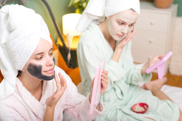 Obraz na płótnie Canvas Two smiling young women in bathrobes and towels putting facial mask on face after shower while sitting in bedroom bed