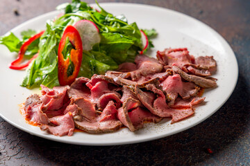 Sliced roast beef with green salad on white plate on dark stone table macro close up