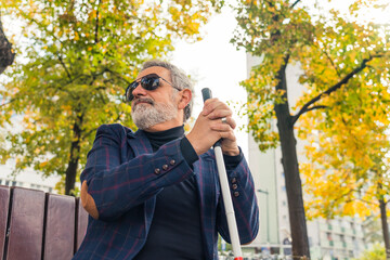 Blind middle-aged man with grey hair and beard in sunglasses sitting on bench in city park holding white cane looking around. Horizontal outdoor shot. High quality photo