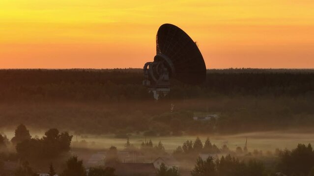 Aerial view. A giant radio telescope for space exploration in a foggy forest during a bloody dawn. Mysterious haze over the forest in early morning.