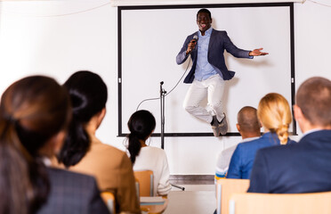 Expressive emotional african american business coach speaking to audience and jumping on stage during motivational training in conference room