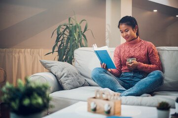 Asian woman siting on the couch at home while reading a book and drinking coffee