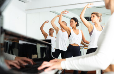 Fototapeta na wymiar Ballet students doing exercises near barre with musician at piano in foreground