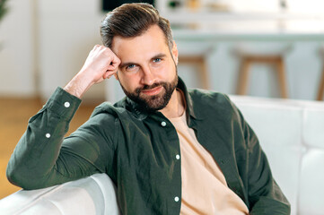 Male portrait. Photo of an attractive positive modern bearded caucasian man, in casual stylish clothes, sits on a sofa in the living room, looks at the camera, smiles friendly