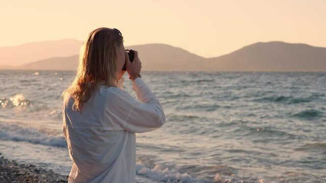 Professional photographer blonde young woman near sea on shore. sunset, unrecognizable girl photographs nature and ocean waves at dawn. traveler captures moments of nature while traveling.