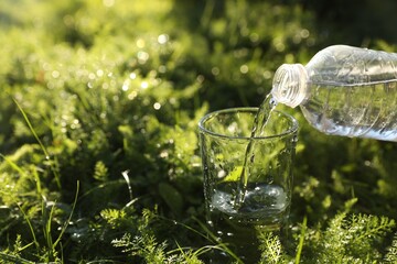 Pouring fresh water from bottle into glass on green grass outdoors. Space for text