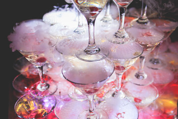Pyramid of glasses with alcoholic beverages, filling and pouring beautiful pyramid line of different colored alcohol cocktails with champagne and dry ice on a party, catering banquet table