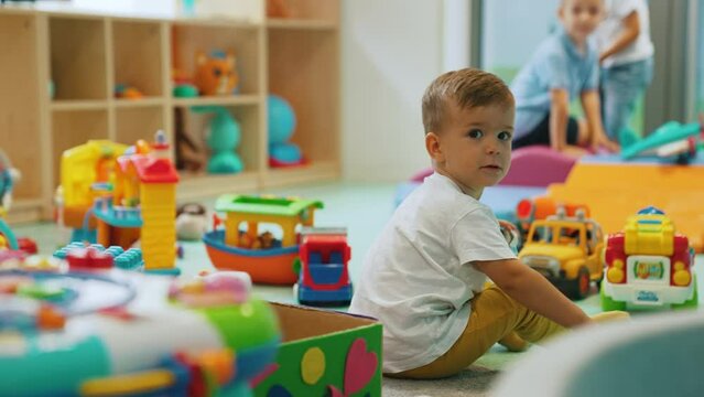 Small white boy playing with toy car in nursery during playtime surrounded by colorful toys and other kids. Multiracial group. Horizontal video . High quality 4k footage