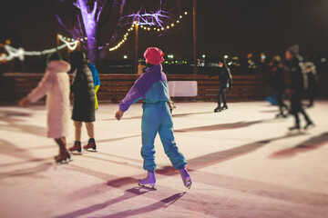Girl ice skating on the ice rink arena with happy people around, concept of ice skating in winter, winter activities, holiday christmas time, with new year decoration and illumination - obrazy, fototapety, plakaty