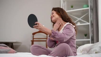 Confident mature woman applying moisturizer on neck skin smiling sitting on bed at home. Wide shot of Caucasian lady in pajamas indoors in the morning. Beauty routine and domestic lifestyle