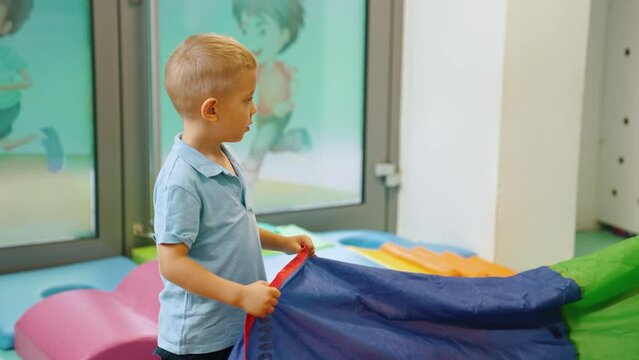 Small blond white boy in nursery holding rainbow play parachute during playtime and looking around. Horizontal indoor video. High quality 4k footage