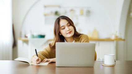 Young asian woman sitting at table and taking notes in notebook. Working on table with computer laptop, document paperwork and cup of coffee. Student learning online