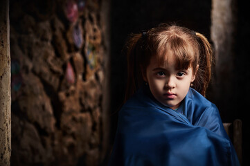 A little girl wrapped in a Ukrainian flag looks at the camera. A child of war. Photo project by Golovchenko Dmytro