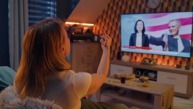 Irritated couple sitting on sofa in living room with snacks, watching politics TV news and speech of President of United States, getting angry, showing middle finger. TV live broadcasting.