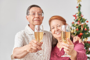 Happy senior latin couple toasting towards camera during Christmas or New Year, celebrating holidays together. Selective focus on the glasses.