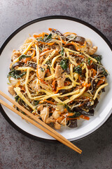 japchae or stir-fried Korean vermicelli noodles with vegetables and pork topped with sesame closeup on the plate on the table. Vertical top view from above