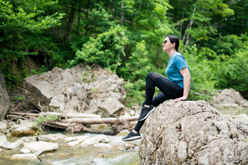 Obraz premium Woman hiker sitting on rock by river in the forest. Female exploring untamed natural areas along the river. Outdoor recreation, active leisure, local getaway concept